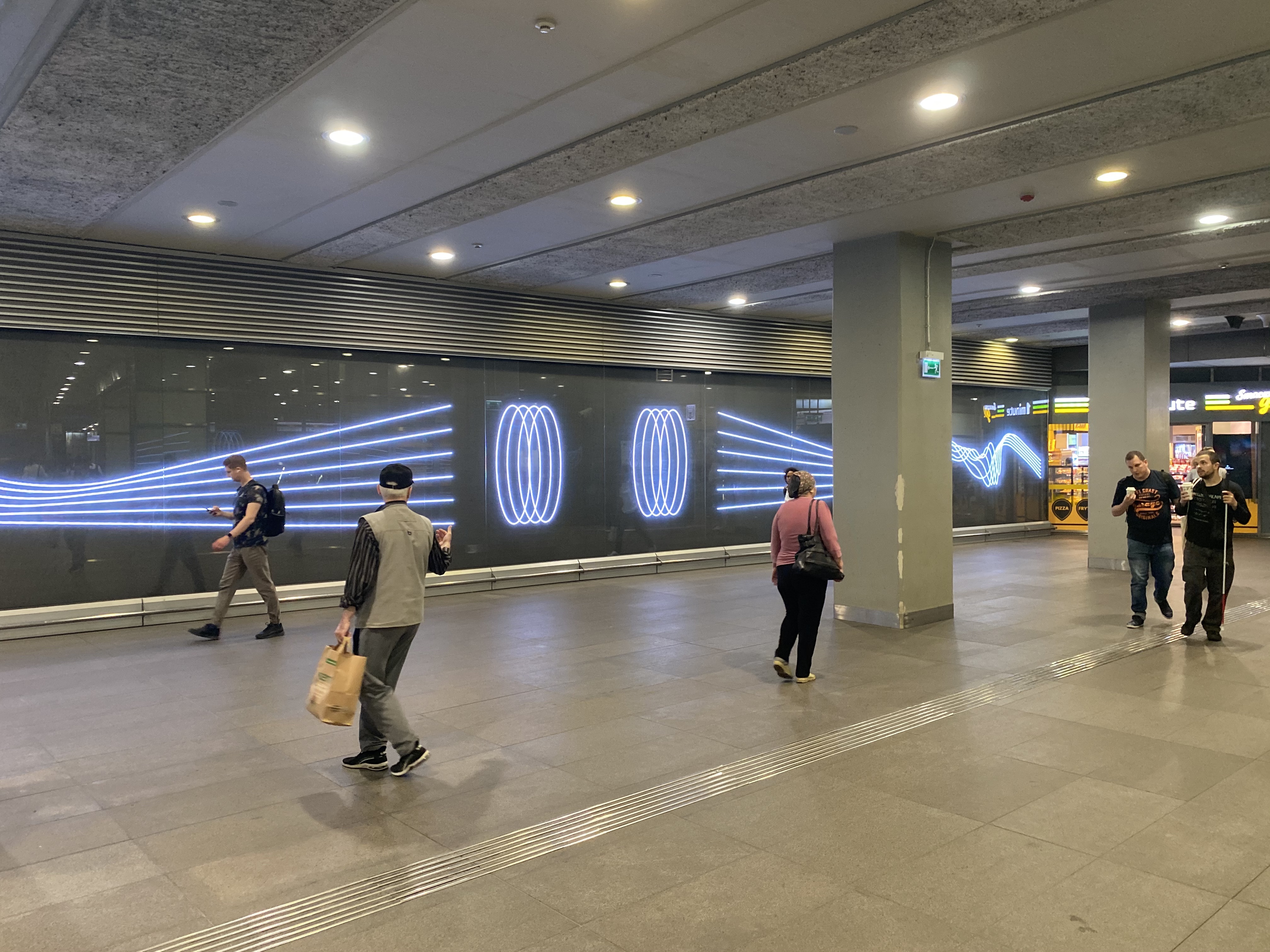 Minimalistyczne grafiki wyświetlone na dużych ekranach LED na stacji metra Świętokrzyska. Widać przechodzących obok pasażerów. 