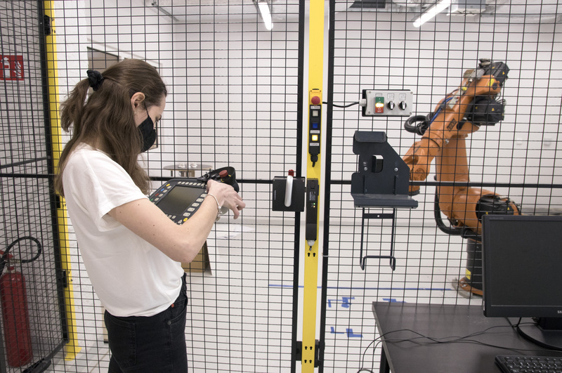 A woman operates a KUKA robot. A black net separates her from the machine for safety.