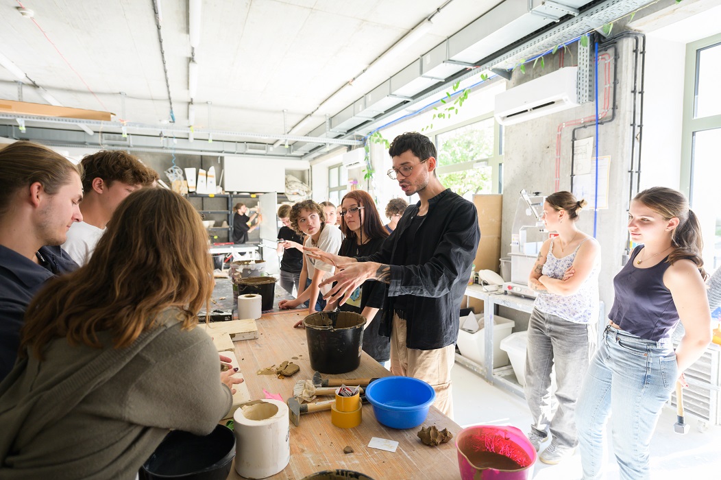 Studenci stoją przy stole roboczym w pracowni ceramiki i słuchają prowadzącego.