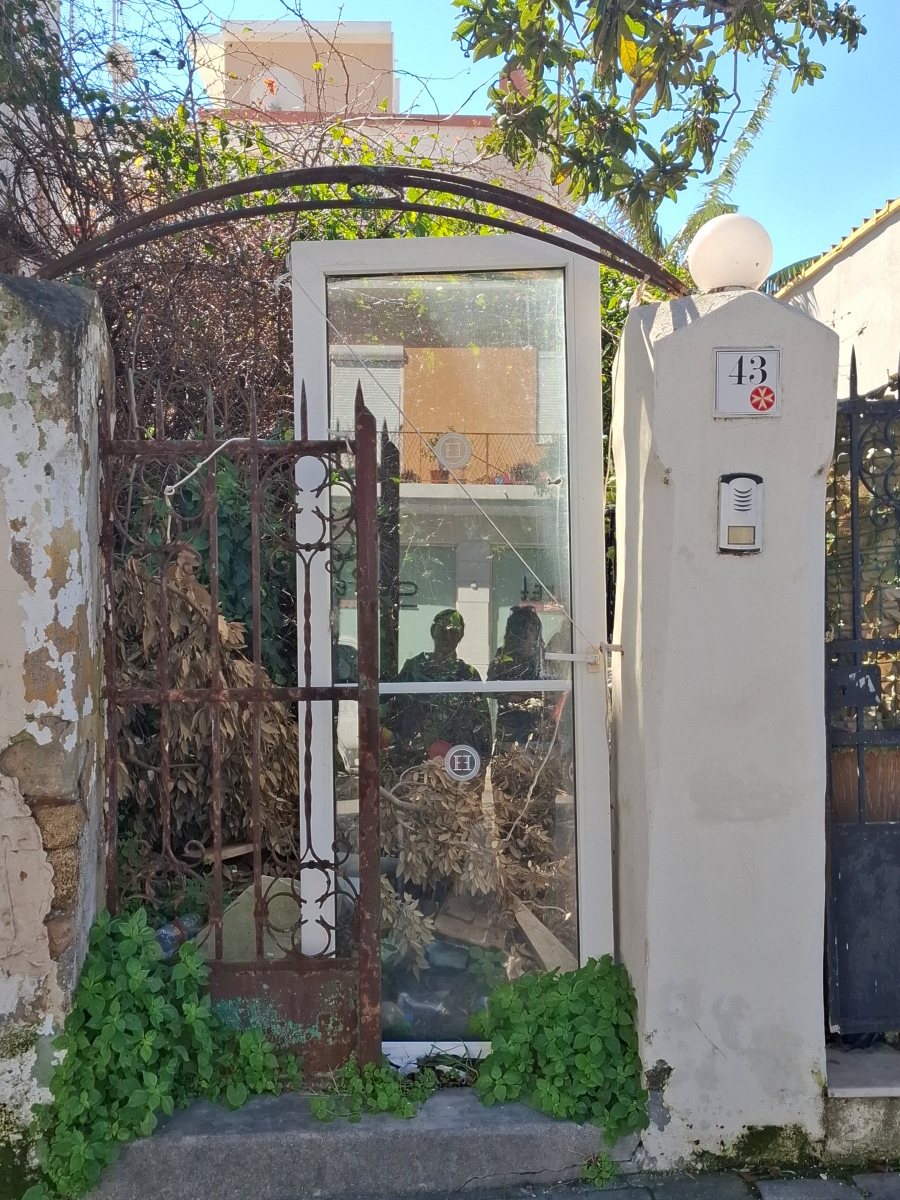 Rdzewiejąca brama z przeszklonymi drzwiami osadzona w jasnym murze z numerem 43. W szybie odbijają się sylwetki osób robiących zdjęcie.