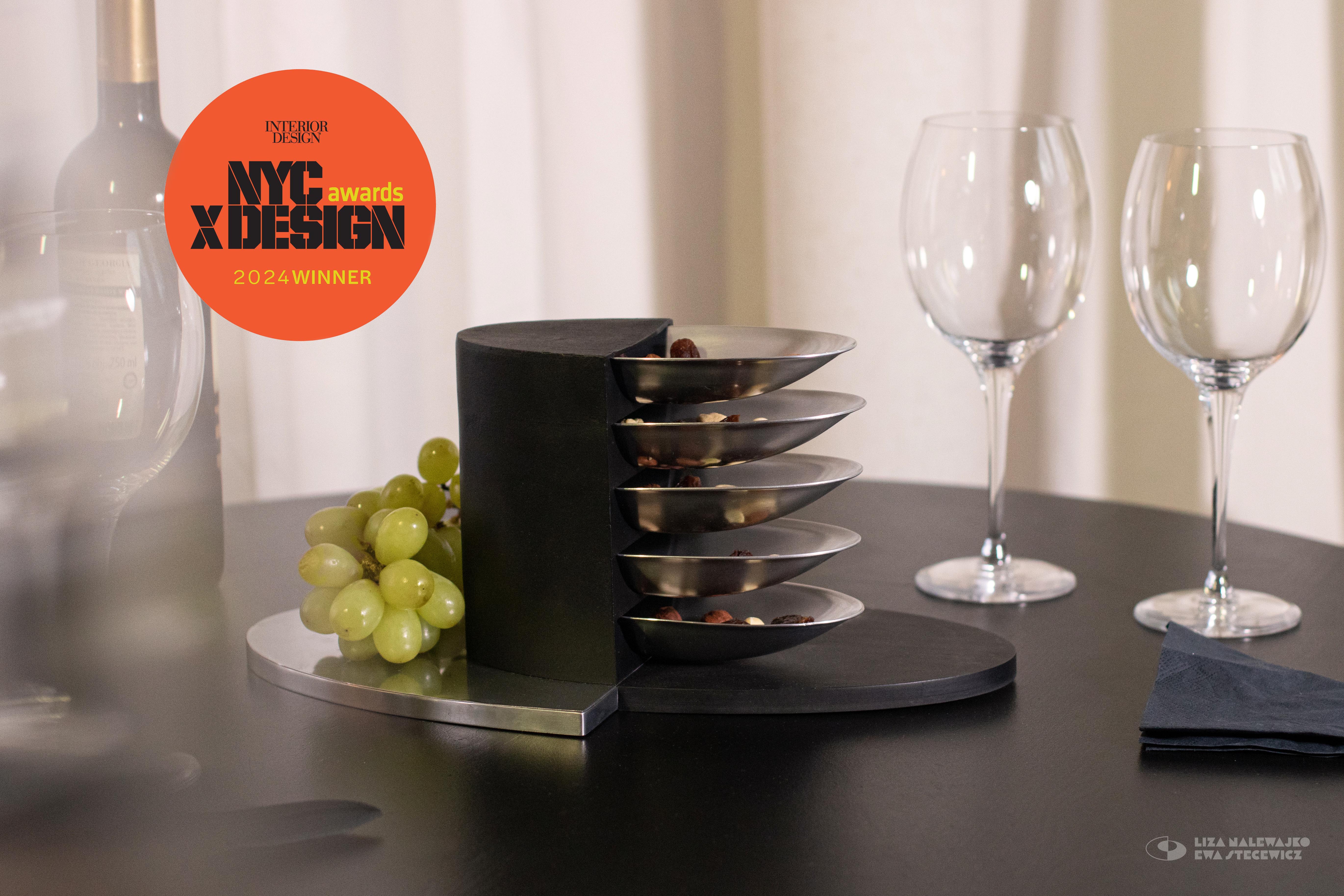 Sześciopoziomowa patera z wyjmowanymi miseczkami postawiona na stole koło kieliszków i kiści winogron. Na zdjęciu znajduje się logo konkursu NYCxDesign Awards 2024.