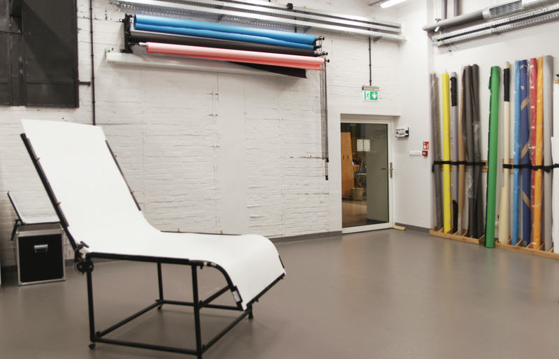 The interior of a photo-video studio. In the foreground is a platform with a white background for photographing small objects. In the background are rolled up photographic backgrounds of different colors leaning against the wall.
