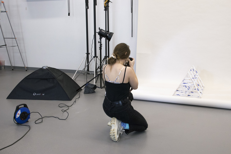 A young girl kneels down and photographs a three-dimensional work set against a white photographic background.