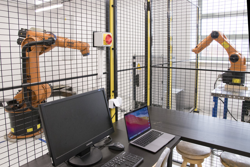 Wnętrze pracowni KUKA. Widok na biurko z komputerem i dwa ramiona robotyczne KUKA oddzielone od biurka czarną siatką.
