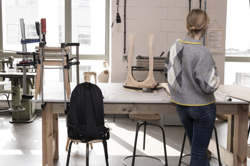 A young girl is working in a Woodworking Workshop. She is standing behind a table with the elements of a wooden seat on it.