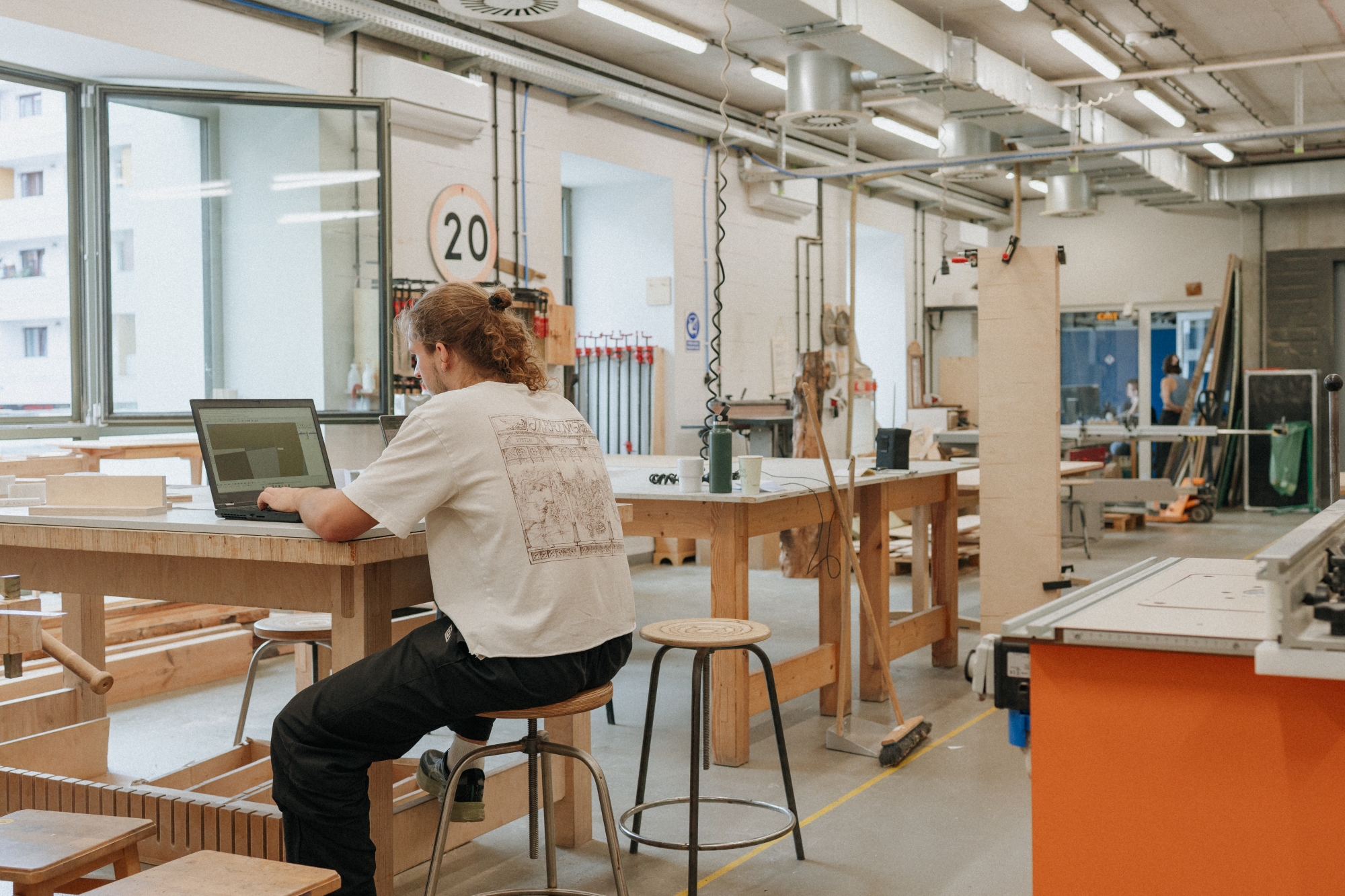 Interior of the woodworking workshop, with a man seated at a worktable working on a computer.