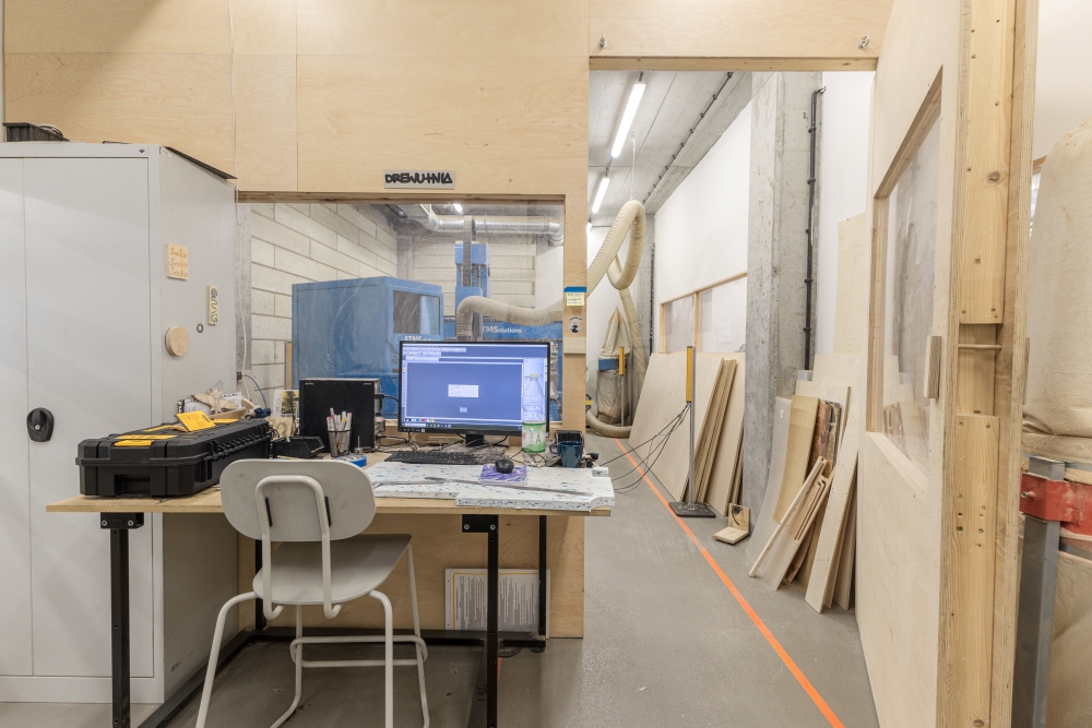 Interior of the woodworking workshop, showing a computer workstation with another room visible behind it.
