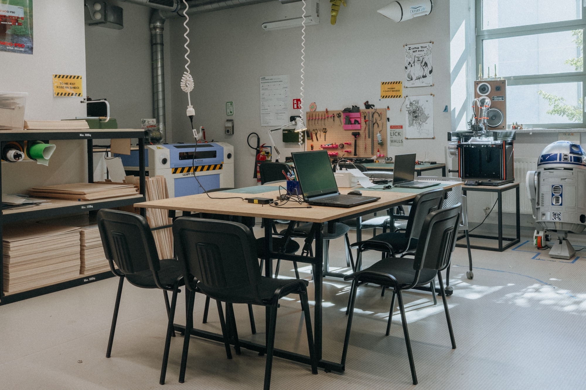 Wnętrze warsztatu digital. Widok na duży stół, krzesła i regały. Po prawej stronie stoi robot R2D2
