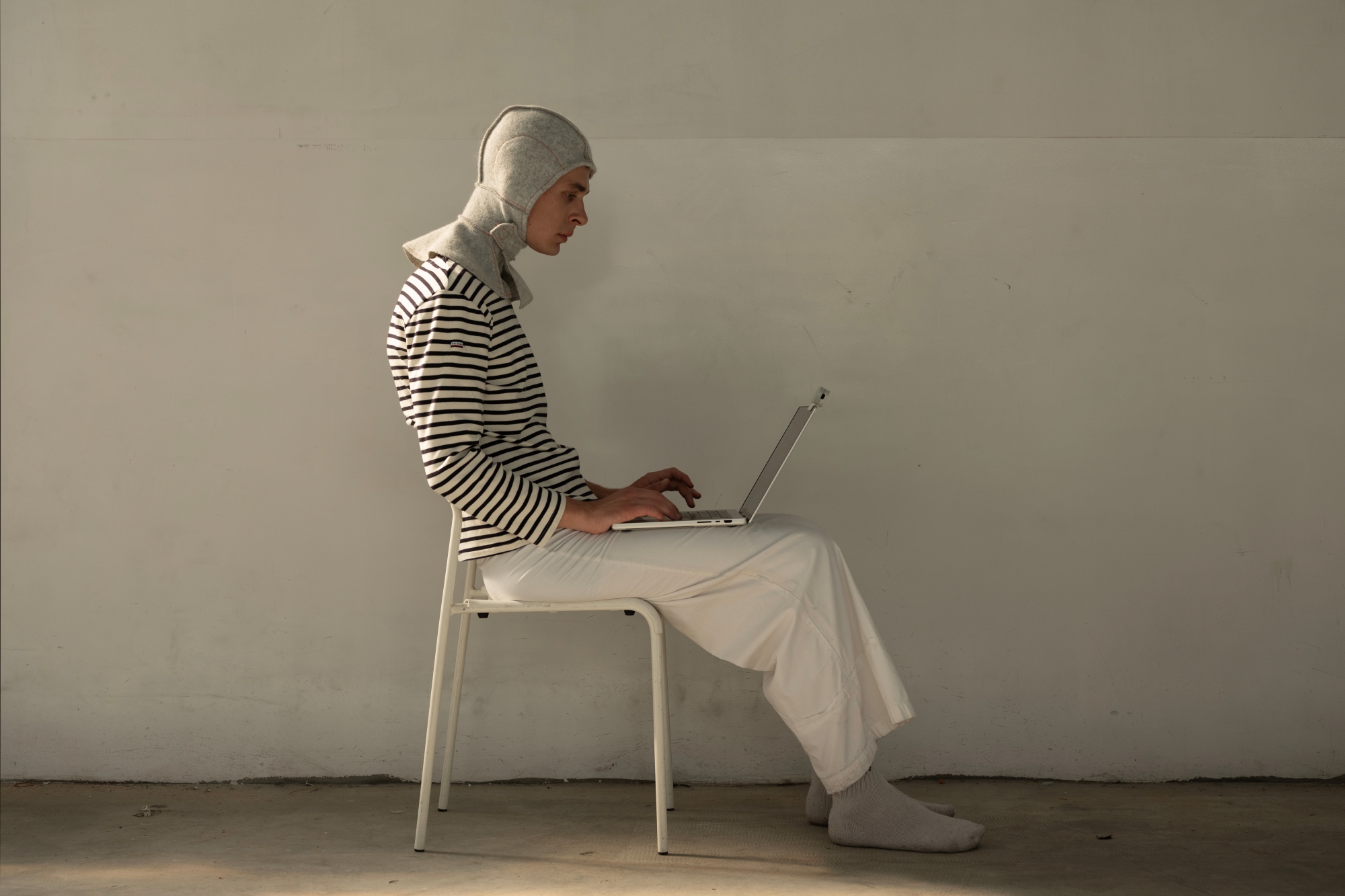 A man wearing a hood designed by Pola Kołodziej sits on a chair with a laptop on his lap.