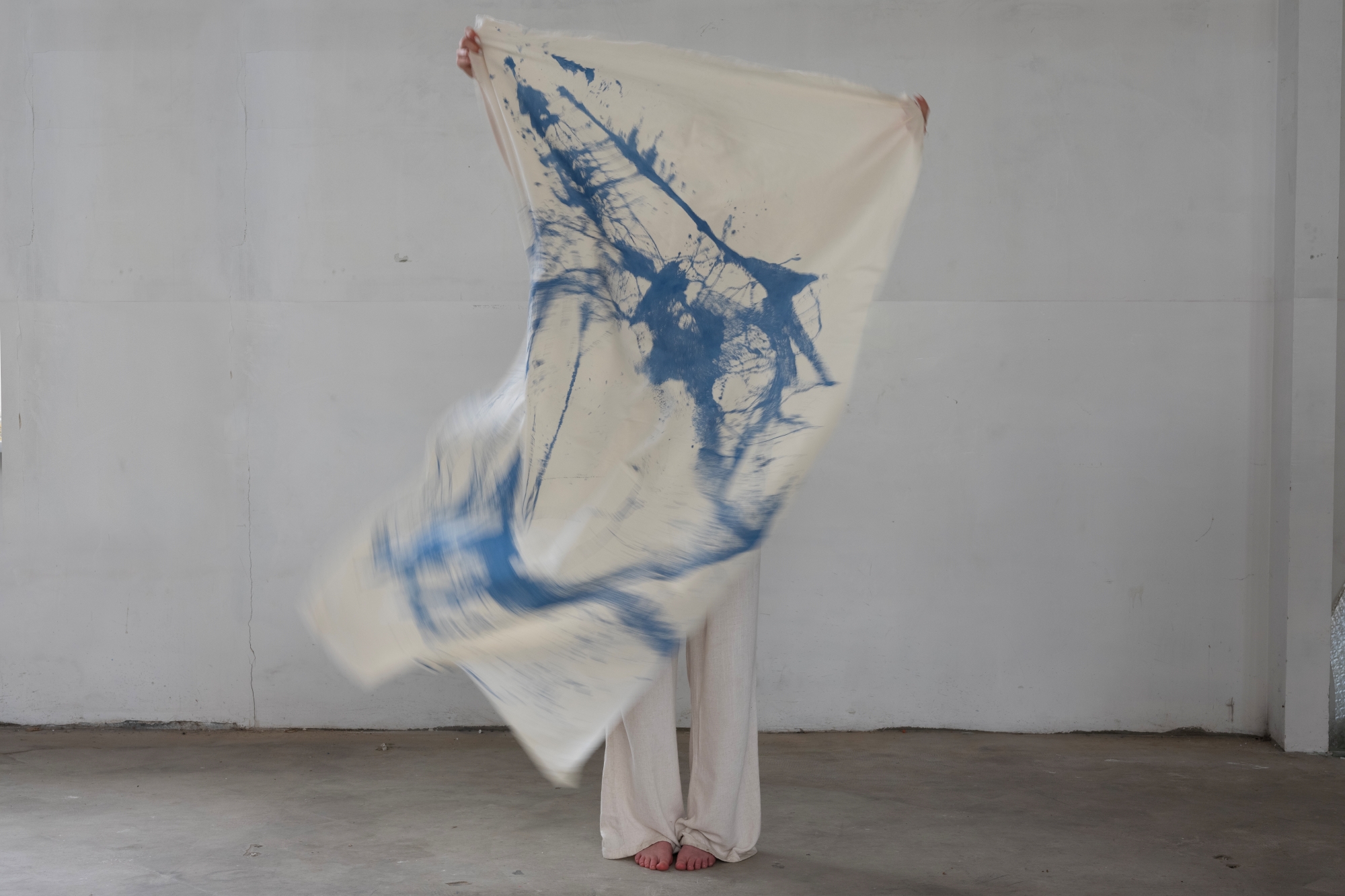 A person is holding up a piece of fluttering fabric with an abstract print. Their legs and hands are visible behind it.