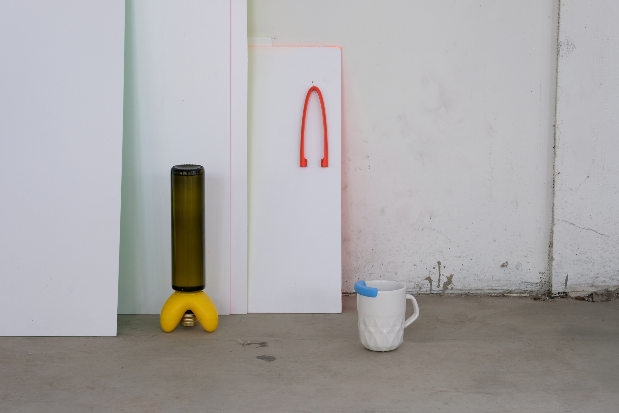 A set of gadgets designed by Przemysław Sokół: a bottle rack, a grabber, and a mug with a seal stand against a concrete wall.