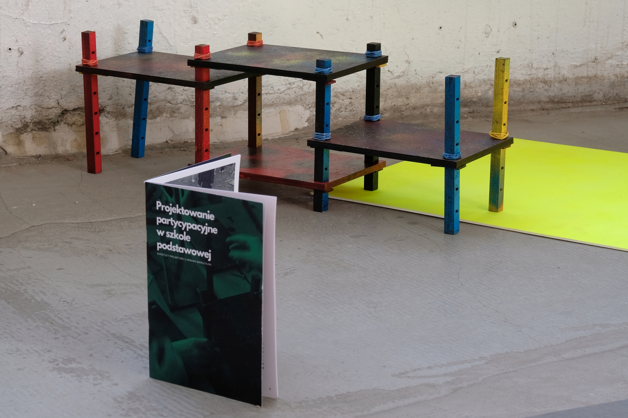 A paper publication lies in the foreground, with a sculptural object resembling a plant stand in the background.