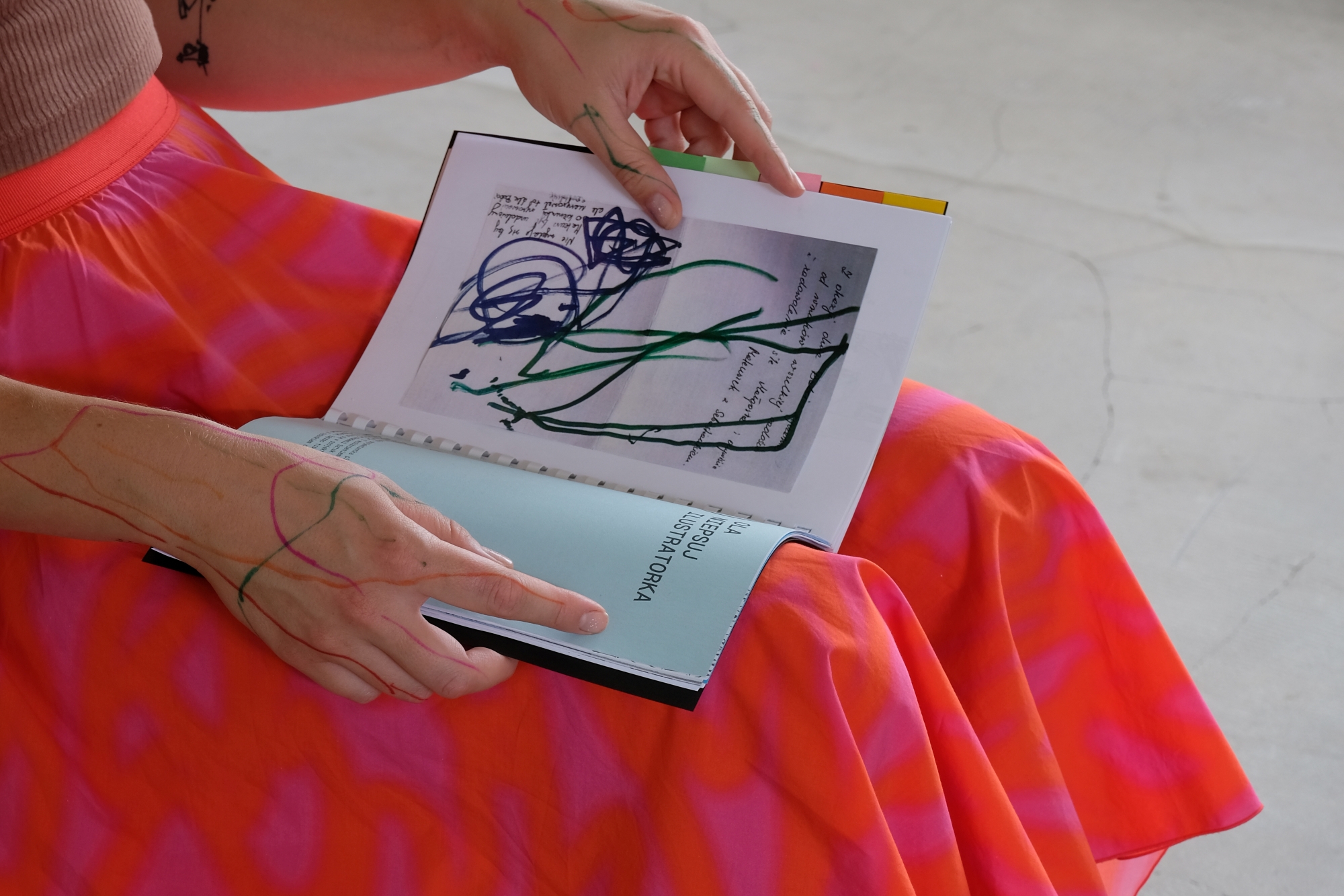 A woman holds a paper publication in her lap. Abstract lines painted with markers adorn her hands.