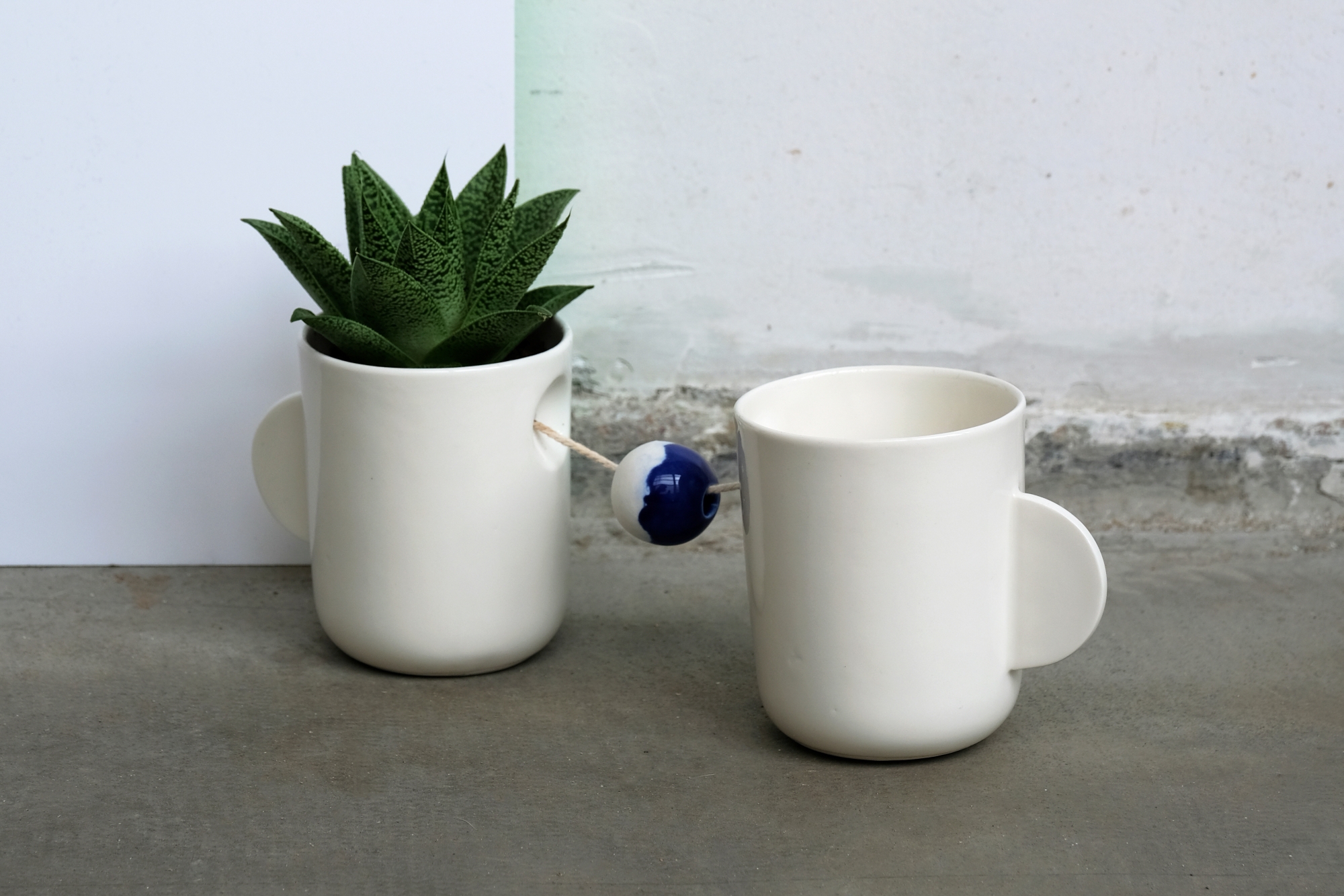 Two ceramic mugs designed by Katarzyna Sadowska; the mugs are connected with a string, and one of them contains a small plant.