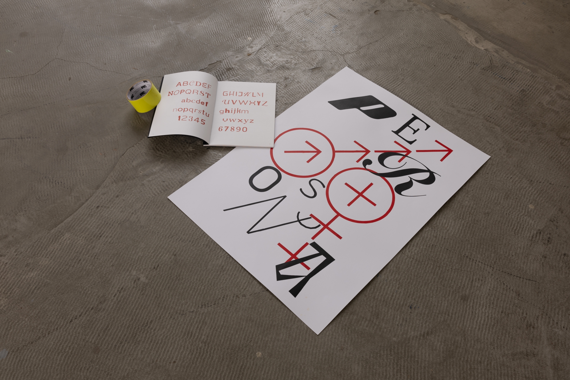 Plakat z czarno-czerwoną kompozycją typograficzną leży na betonowej podłodze obok otwartego zina ze wzornikiem krojów pisma i rolki żółtej taśmy.