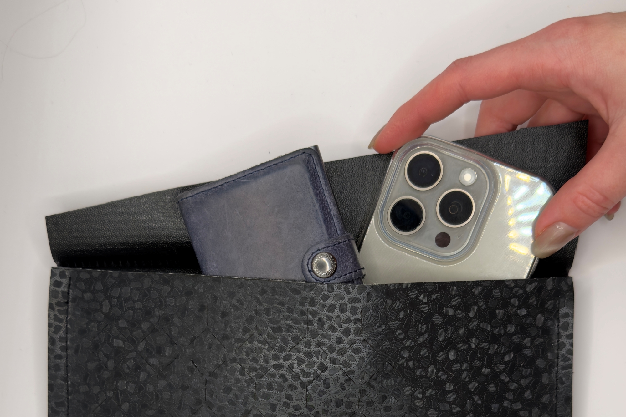 A hand puts a smartphone into a bag made of roofing membrane. There is already a wallet in the bag.