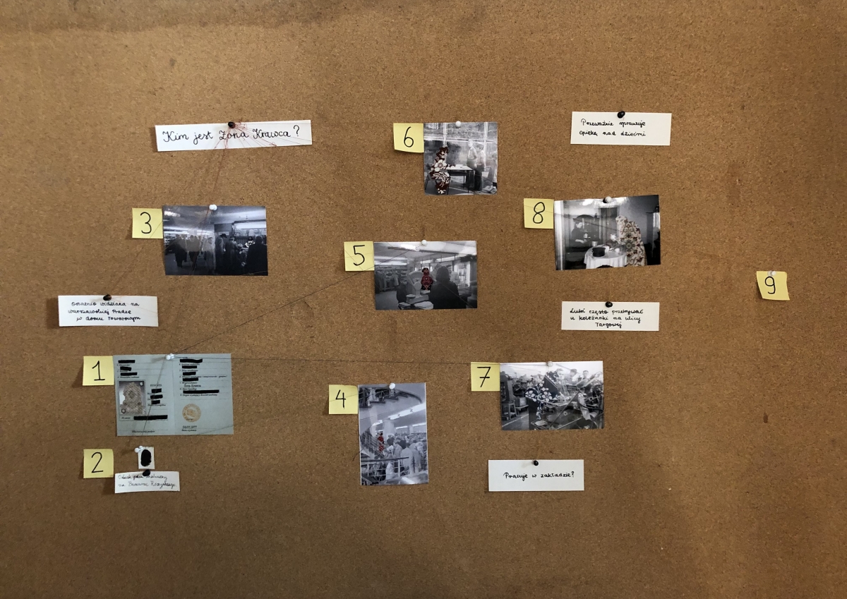 Tablica korkowa przypominająca tablicę detektywistyczną. Przypięto do niej fotografie, zamazany dokument tożsamości i odręczne notatki, które połączono nicią. Na najwyższej kartce widnieje główne pytanie: 
